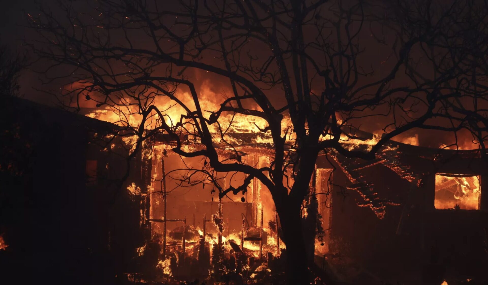 A large house engulfed in flames at night, with a dark, leafless tree silhouetted in the foreground. The bright, intense fire illuminates the structure, casting dramatic shadows and highlighting the trees barren branches.